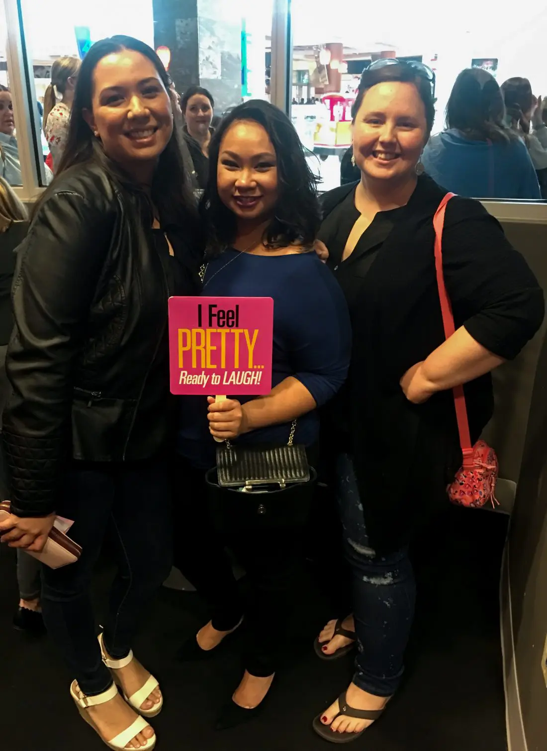Three women holding a I Feel Pretty Film sign. The I Feel Pretty Film is in theaters everywhere today.