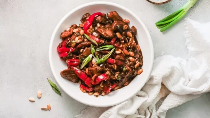 Better than Chinese takeout, make this Kung Pao Chicken at home. A horizonal shot of the Kung Pao Chicken dish being served in a white bowl next to a white napkin.